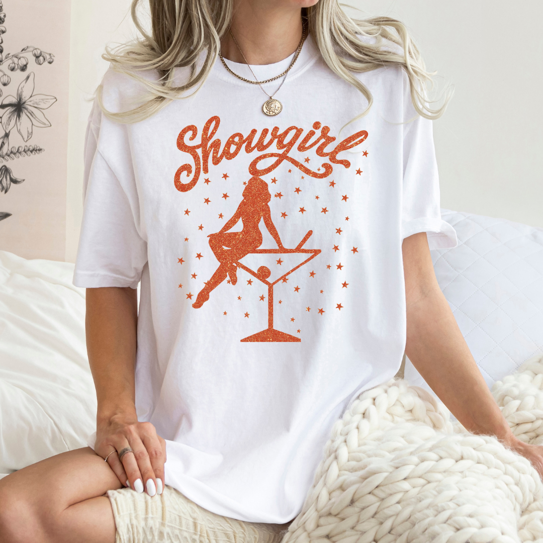 Person wearing a white t-shirt with 'Showgirl' design sitting on a bed.