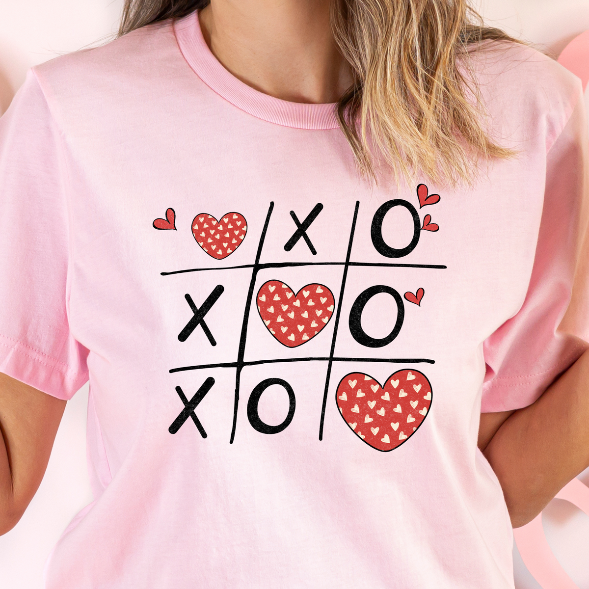 A person wearing a pink t-shirt with a graphic of a 'Naughts and Crosses' game board, filled with red hearts and the text 'LOLs OF COLOURS & SIZES'.
