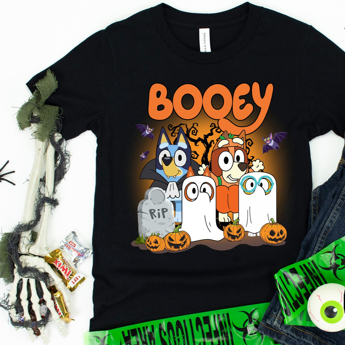 A black t-shirt with a graphic print of the Bluey family in Halloween costumes, accompanied by a pair of sneakers and a 'BOOEY' text graphic. The background has a Halloween theme with decorations and a skeleton.