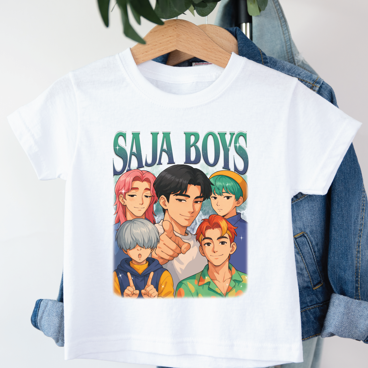 White t-shirt with 'SAJA BOYS' graphic design featuring five animated characters, hanging on a wooden hanger against a denim background.