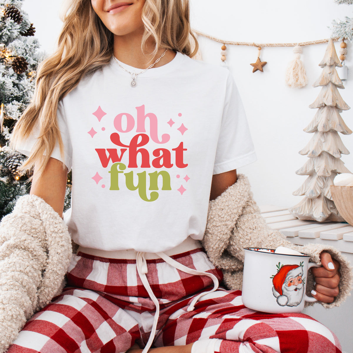 A person wearing a white t-shirt with the text 'Oh what FUN' printed in pink and green colors, sitting in a cozy room with a Christmas tree and gifts in the background.