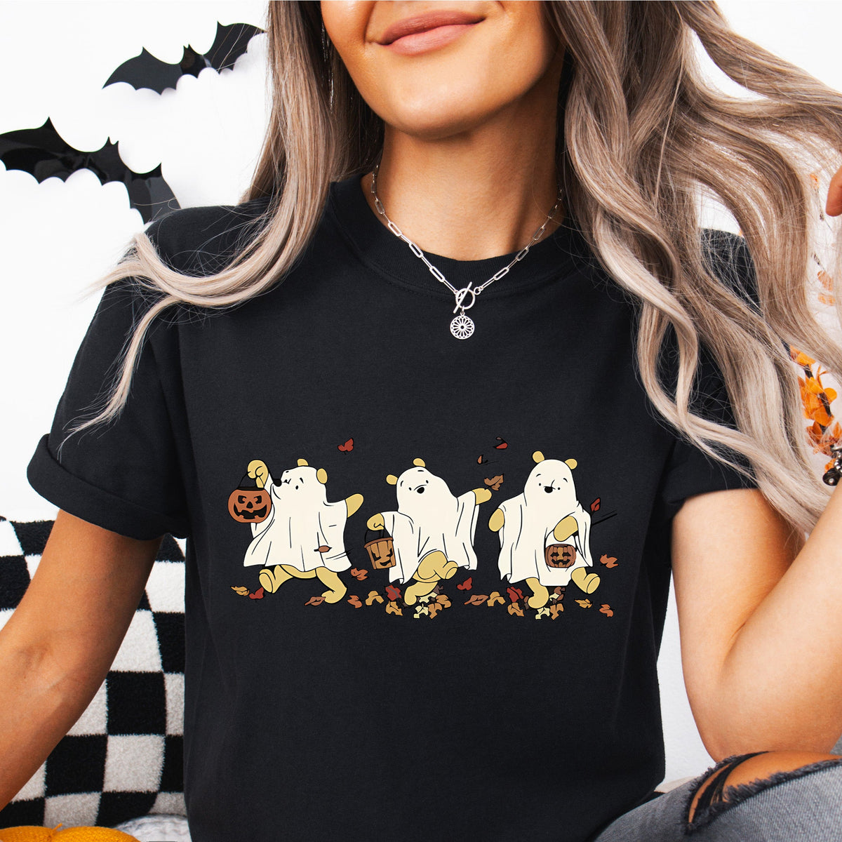 A woman sitting and wearing a black T-shirt with a Winnie the Pooh Halloween design showing three cartoon characters of Pooh in different poses with a pumpkin and checkered background.