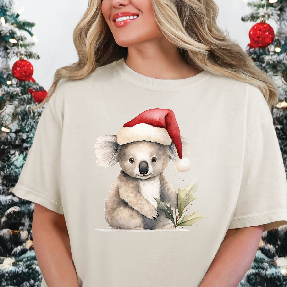 A person wearing a beige t-shirt with a graphic of a koala wearing a Santa hat, and text indicating the t-shirt size and color availability.