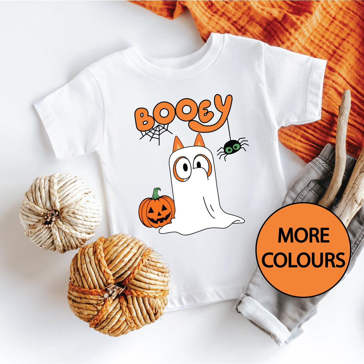 A white t-shirt with a colorful Halloween themed design featuring a ghost, pumpkin, and spider, positioned on a flat surface with orange textiles and a 'MORE COLORS' sign in the background.