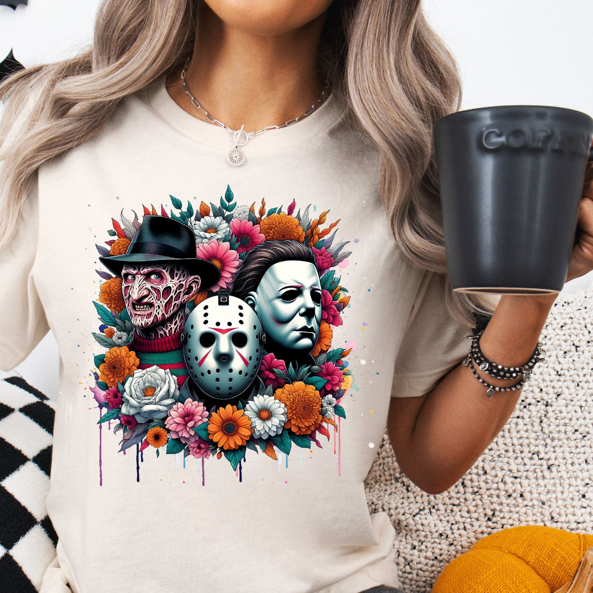 A person wearing a beige t-shirt with a colorful horror-themed design featuring Freddy Krueger, Jason Voorhees, and Michael Myers surrounded by flowers, holding a coffee cup, and seated on a patterned surface.