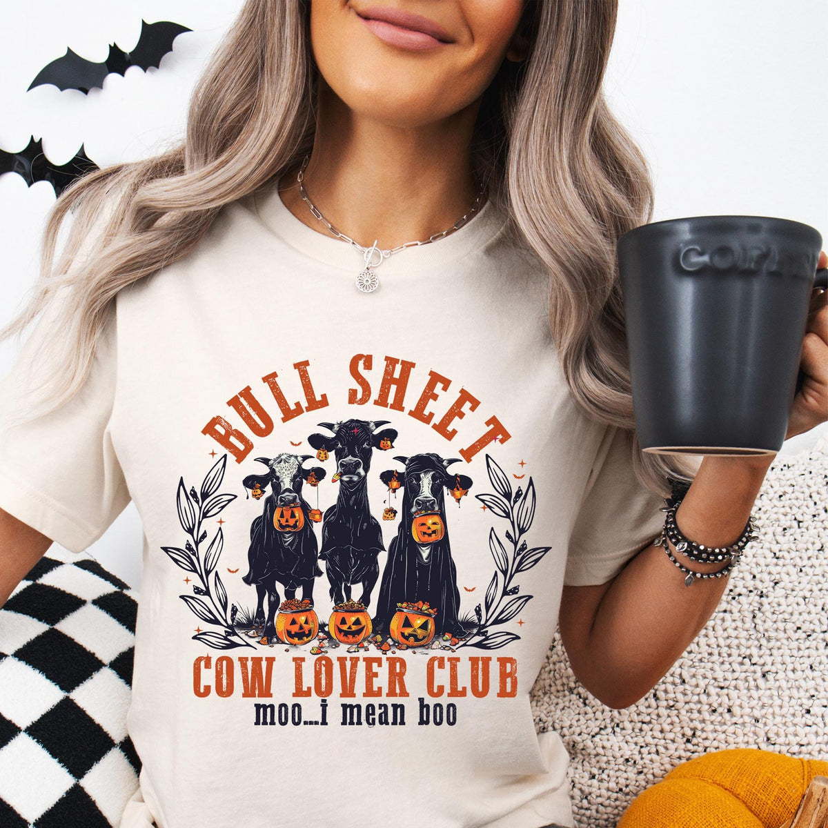 Person wearing a 'Bull Sheet Cow Lover Club' t-shirt with Halloween design, holding a mug.