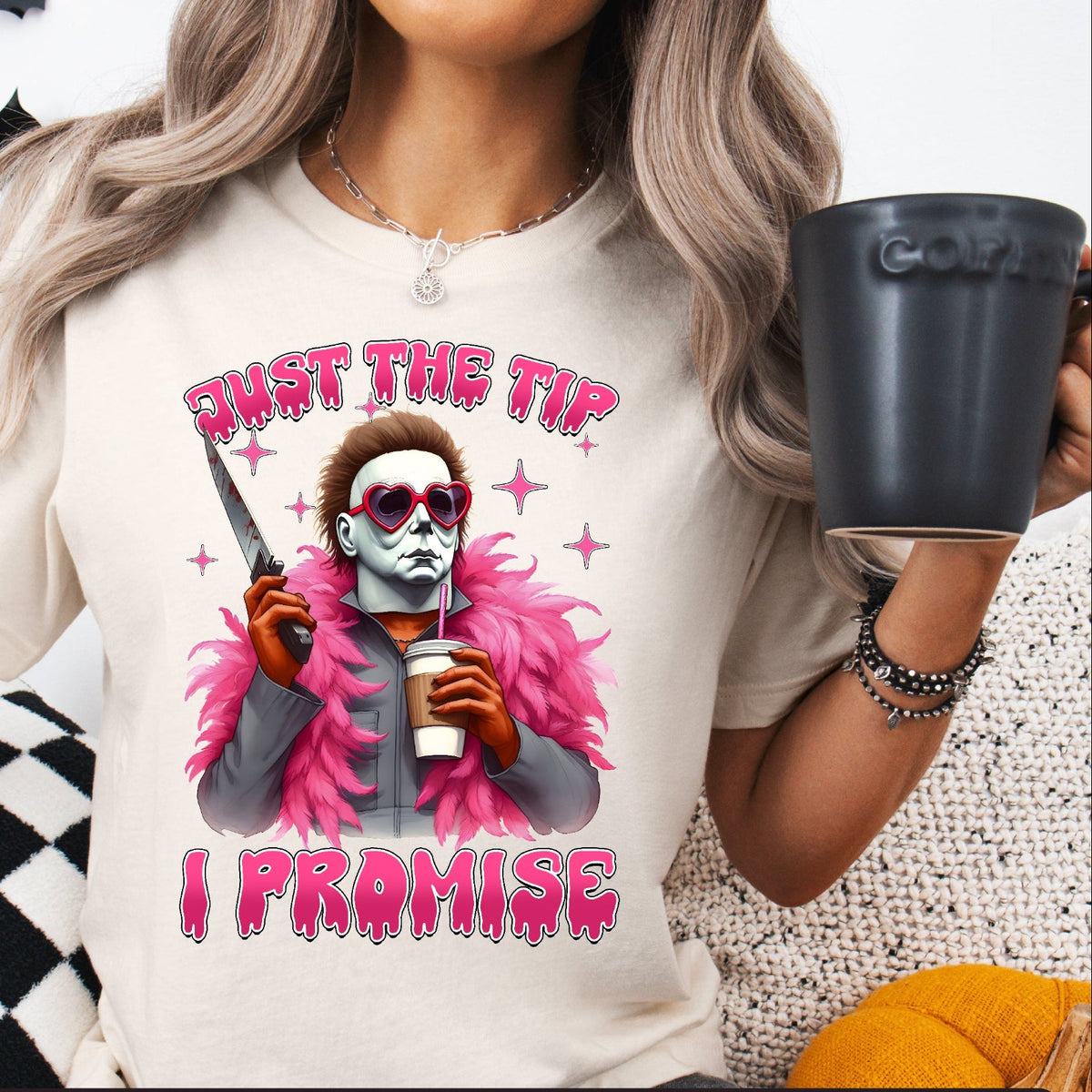 A person wearing a white T-shirt with a graphic print of Michael Myers from Halloween, holding a coffee cup, and text 'JUST THE TIP, I PROMISE'.