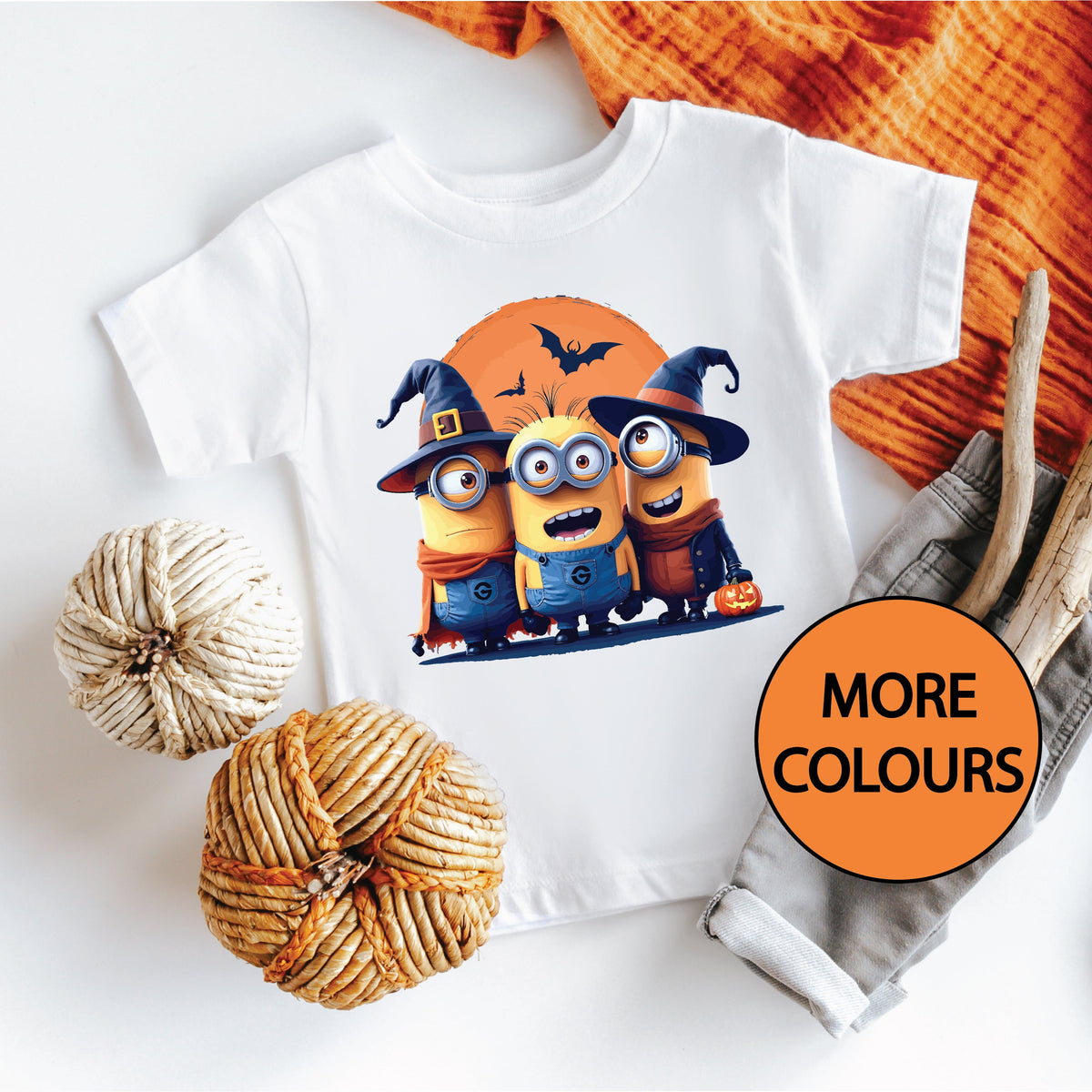 A white t-shirt with a printed design featuring Minions characters in a Halloween setting, displayed on a flat surface with orange textiles and a wooden element in the background.