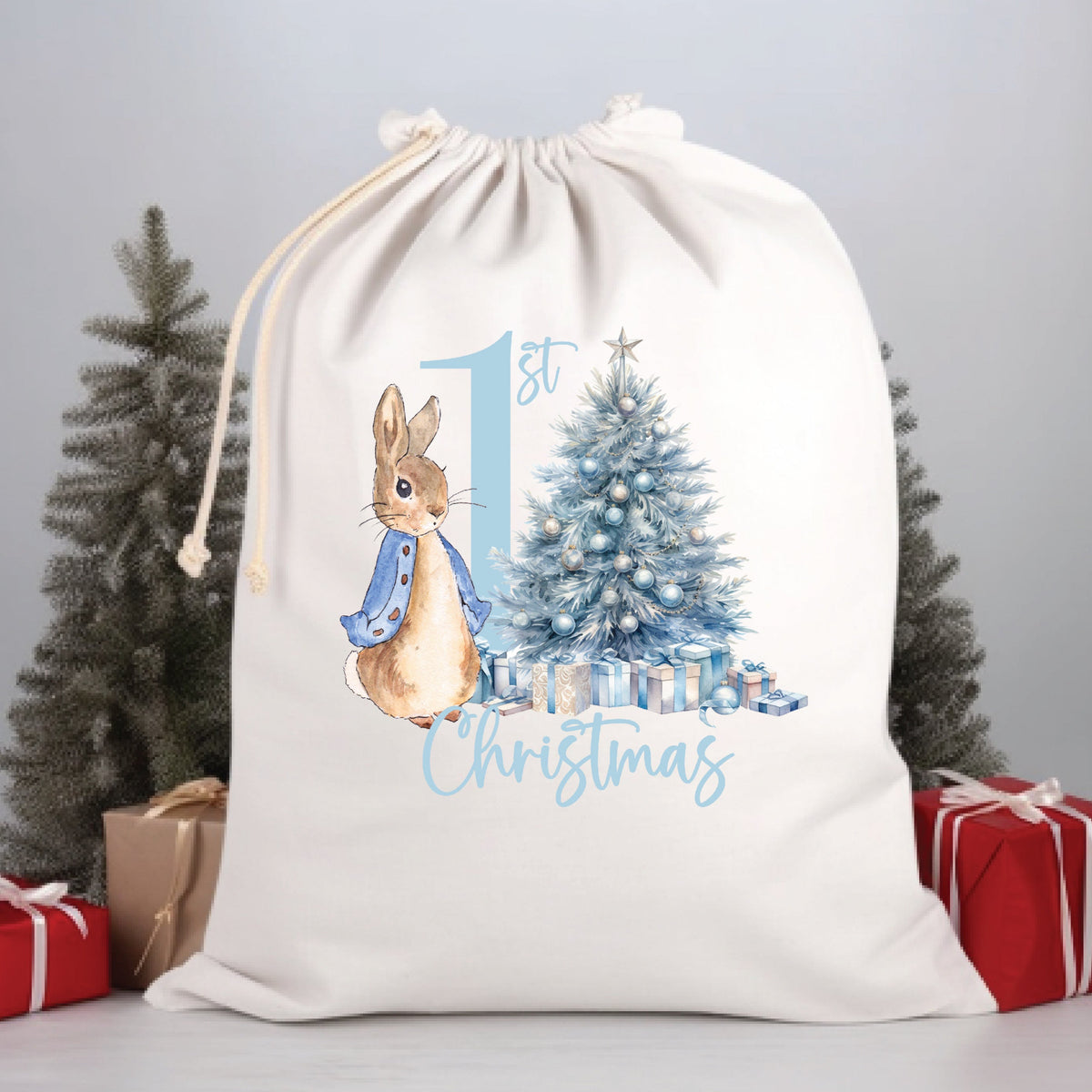 A white Santa sack with a illustration of Peter Rabbit, a Christmas tree, and the text '1st Christmas'. The sack is placed against a grey background with two red gift boxes and a Christmas tree to the sides.