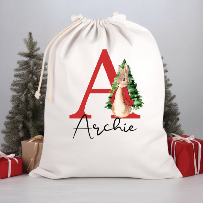 A white Santa sack with a red initial and the name 'Archie', featuring an illustration of Peter Rabbit, presented alongside Christmas trees and presents.
