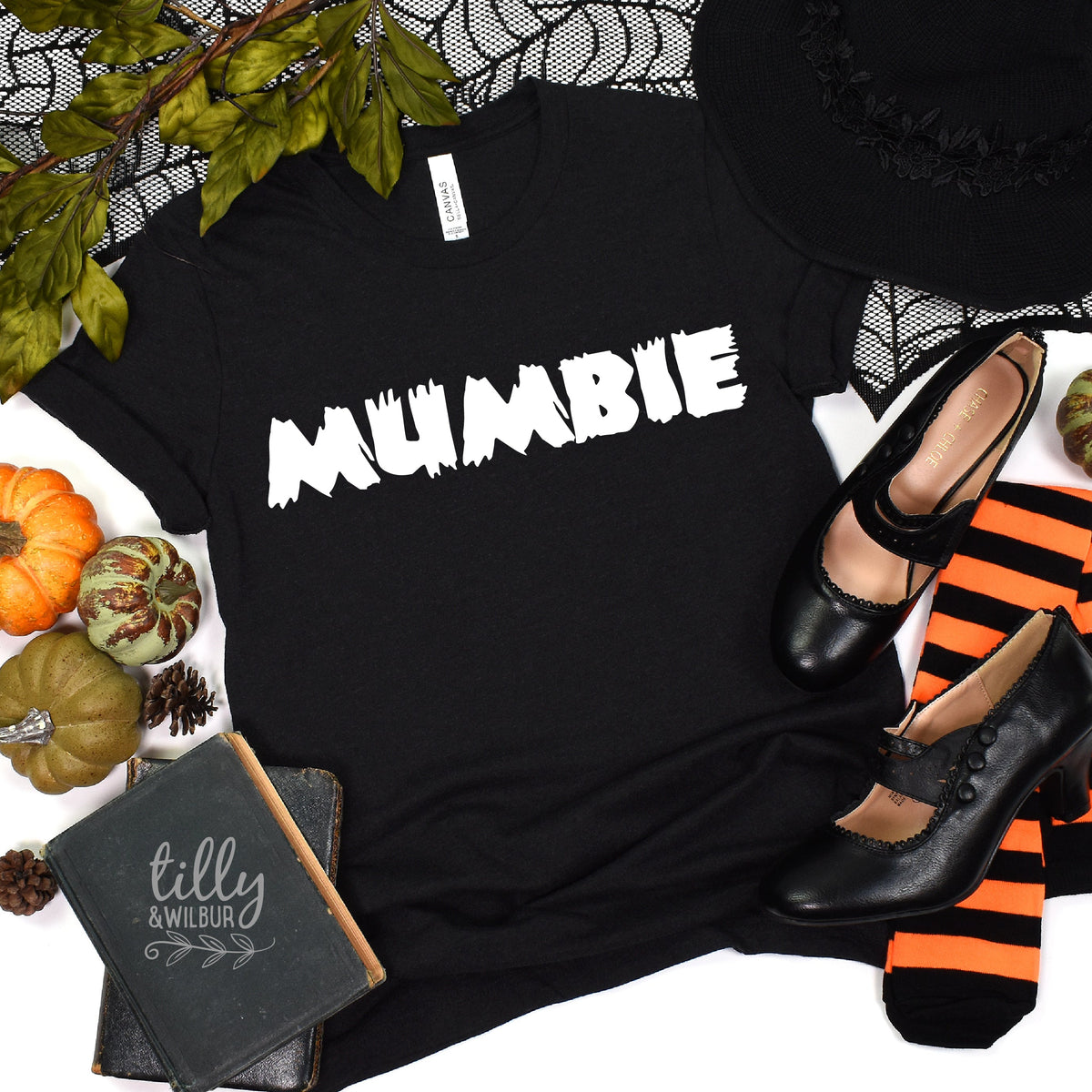 A black t-shirt with the text 'MUMBIE' in white font, displayed flat with a small pumpkin, black shoes, and Halloween-themed accessories around it.