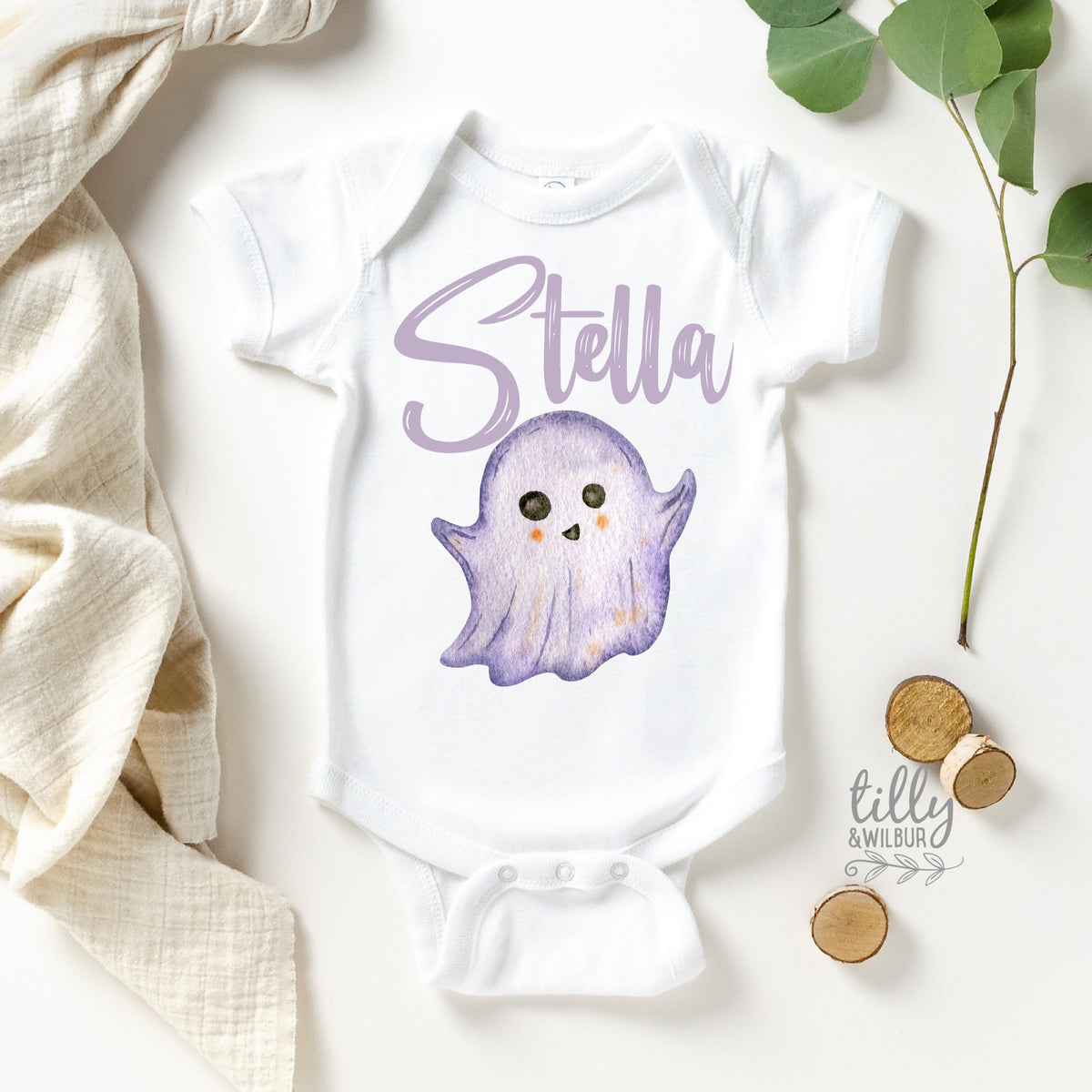 A white baby bodysuit with a custom ghost design and the name 'Stella' printed on the front, accompanied by some leaves and a coin on a white fabric.