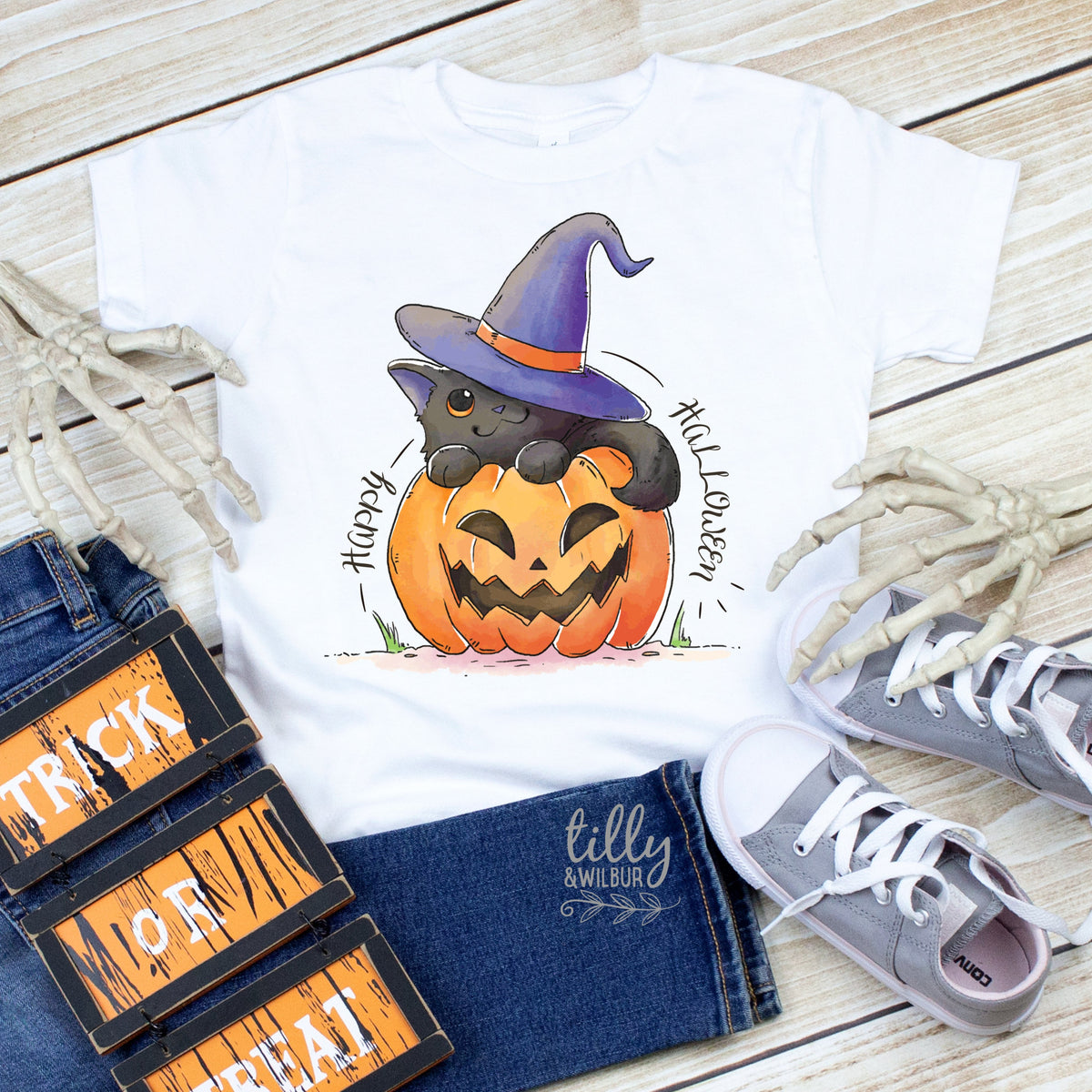 A white t-shirt with a graphic print of a black cat on a pumpkin, wearing a witch hat, displayed with Halloween-themed accessories including a pumpkin, a witch's hat, and a pair of shoes.