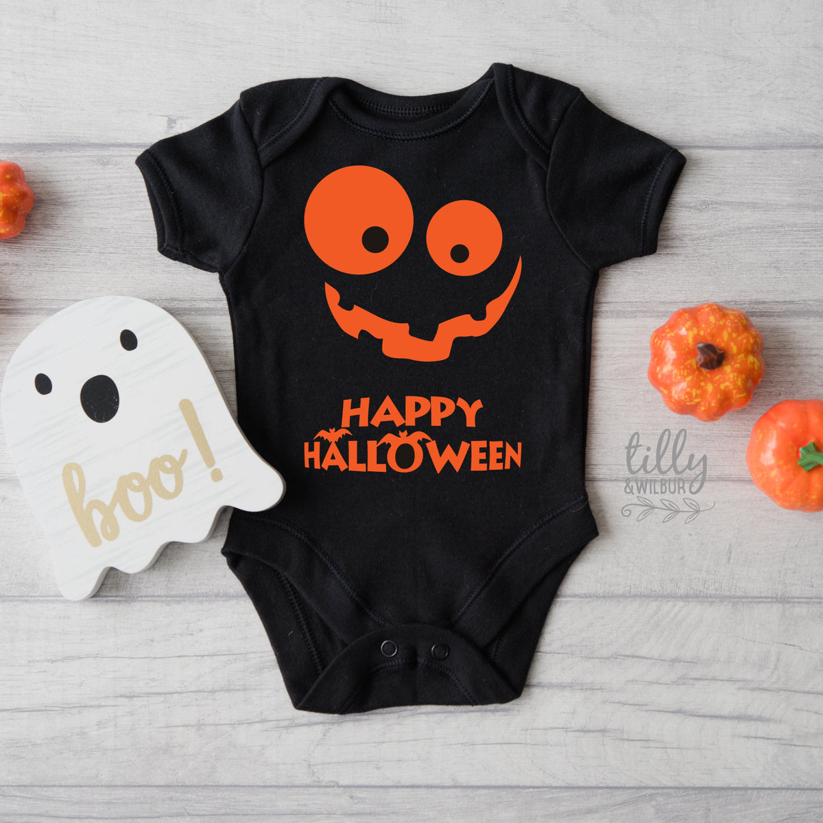 A black bodysuit for babies with a Halloween theme, featuring a large pumpkin face design with the text 'Happy Halloween' and the word 'BOO!' on a wooden block sign, accompanied by real pumpkins and a 'Trick or Treat' sign.