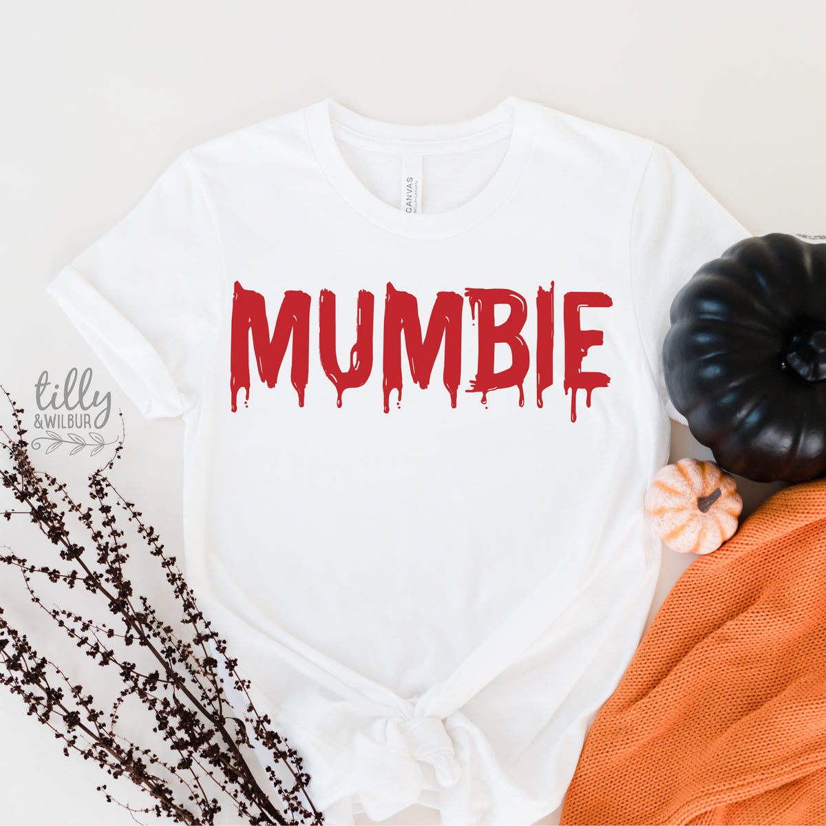 White t-shirt with the word 'MUMBIE' printed in red, accompanied by a pumpkin and decorative items in the background.