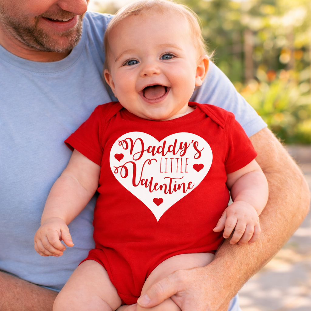 Baby wearing a red 'Daddy's Little Valentine' onesie being held by an adult outdoors.