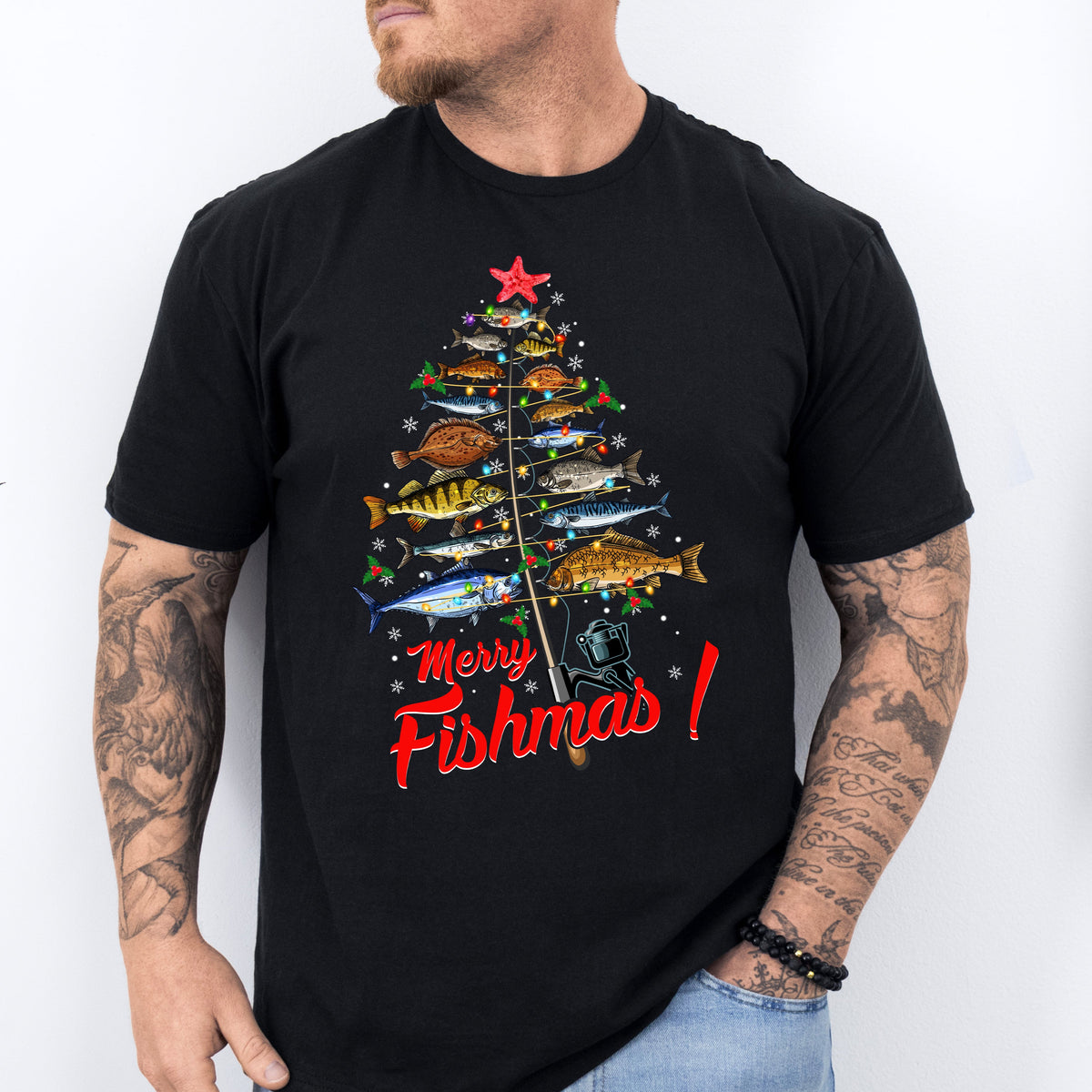 A man wearing a black T-shirt with a colorful Christmas tree design and the text 'Merry Fishmas!' on it, standing in a casual outfit with a tattoo visible on his arm.