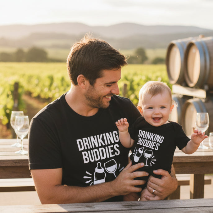 Man holding a baby both wearing black t-shirts with 'DRINKING BUDDIES' text in a vineyard setting.