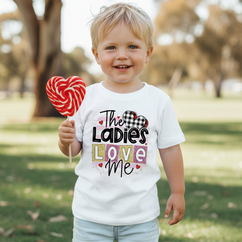 A white baby bodysuit with the text 'THE LADIES LOVE ME!' printed in black, surrounded by red and pink hearts, displayed next to a pair of blue jeans.