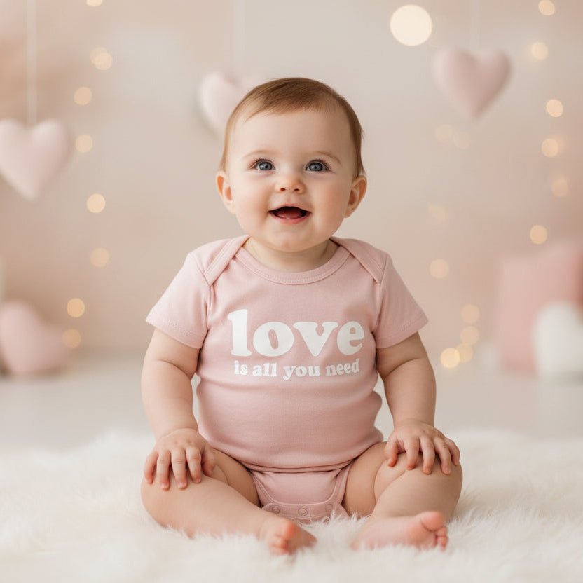 Baby wearing a pink 'love is all you need' onesie in a softly lit room with heart decorations.
